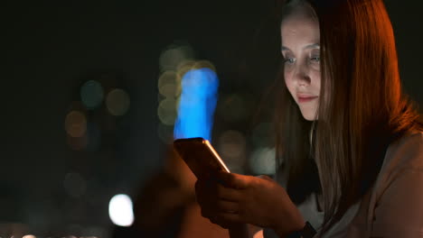 At-night-a-young-girl-sits-on-the-grass-in-a-big-city-and-looks-into-the-smartphone-screen-writes-a-message-and-maintains-her-personal-blog.